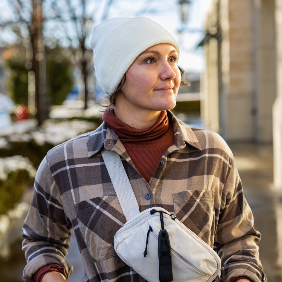 Photograph of a woman wearing a white beanie, brown and beige plaid shirt, and an off-white fanny pack in an outdoor urban setting with subtle snow and muted background architecture.