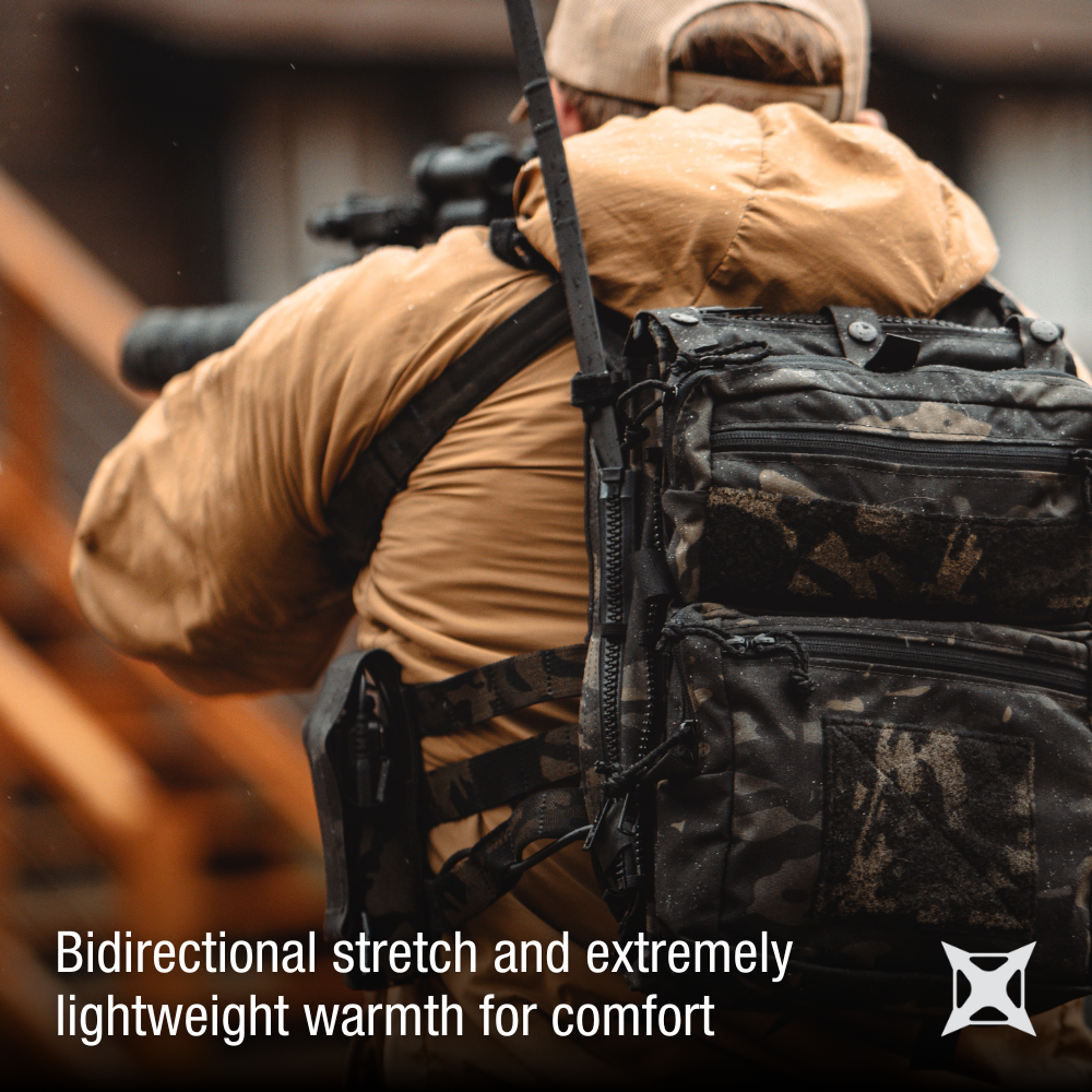 Photograph of a person wearing tan outerwear and a tactical backpack with black and tan camouflage near a wooden structure showing black zippers and multiple pockets