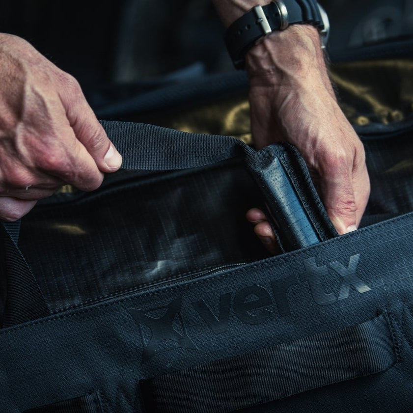 Photograph of a person's hands packing a black Vertix duffel bag featuring robust straps and a textured interior showing a dark blue item inside