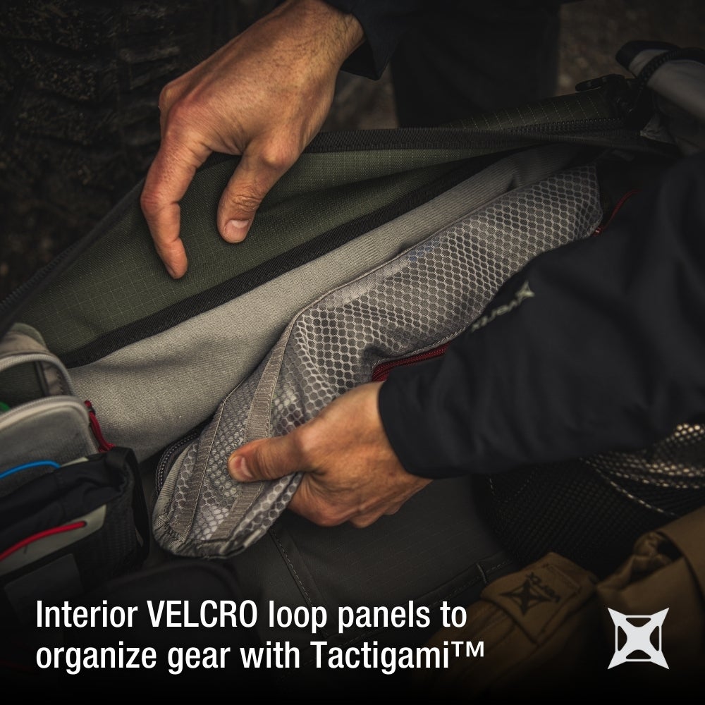 Product photography showing a person's hands arranging grey mesh and olive green Velcro interior panels inside a Tactigami bag featuring red zipper pulls and a textured fabric