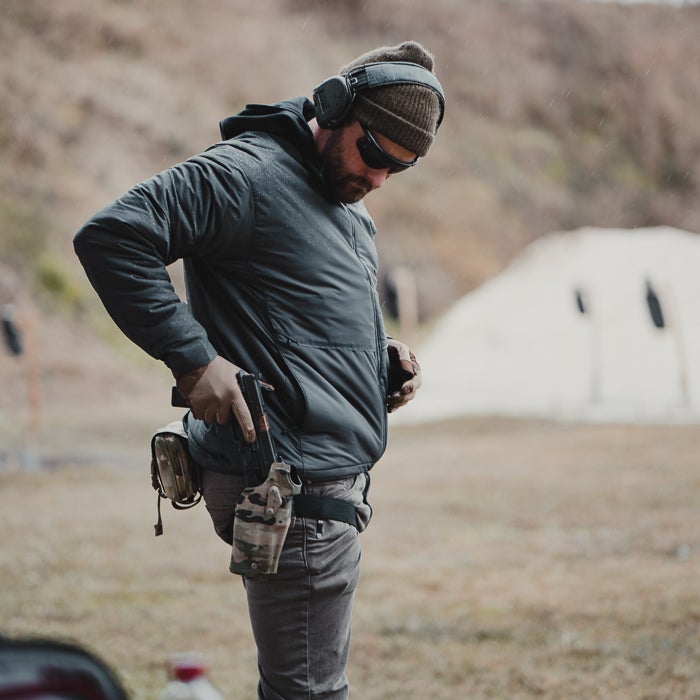 Photograph of a man at an outdoor shooting range adjusting his handgun holster; he's wearing a dark jacket, brown knit hat, and camouflage holster, with a target backdrop visible in the background