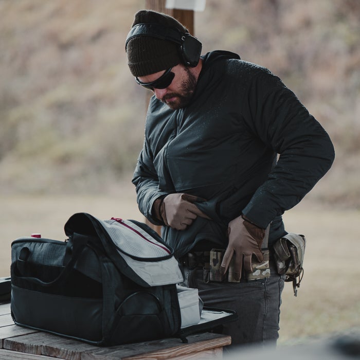 Photograph a man at an outdoor shooting range adjusting his firearm near a gray and black range bag wearing gloves earmuffs and sunglasses