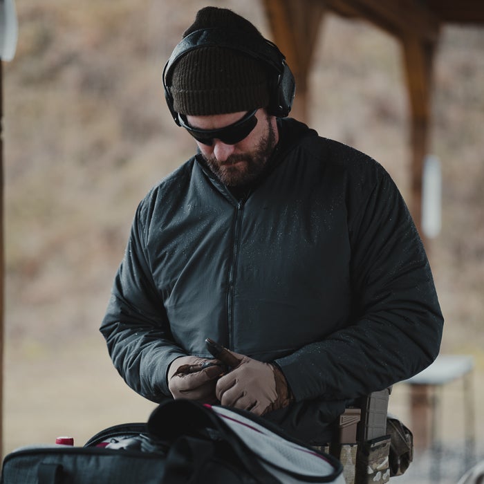 Photograph of a man loading ammunition at an outdoor shooting range wearing a dark jacket tan gloves and noise canceling headphones