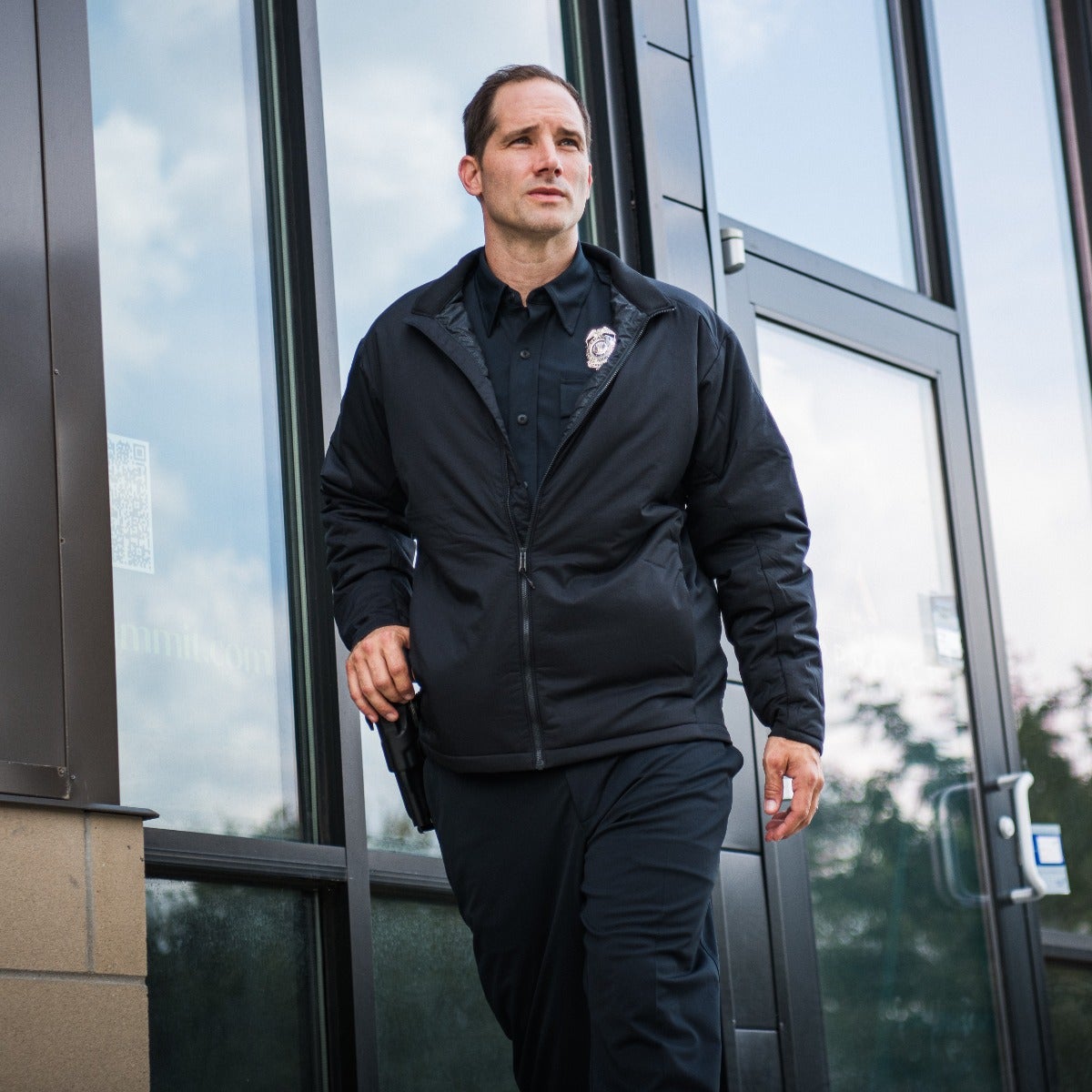 Photograph of a police officer in a black jacket and pants walking near a modern building with large glass windows and dark metal framing showcasing a silver badge and a holstered gun