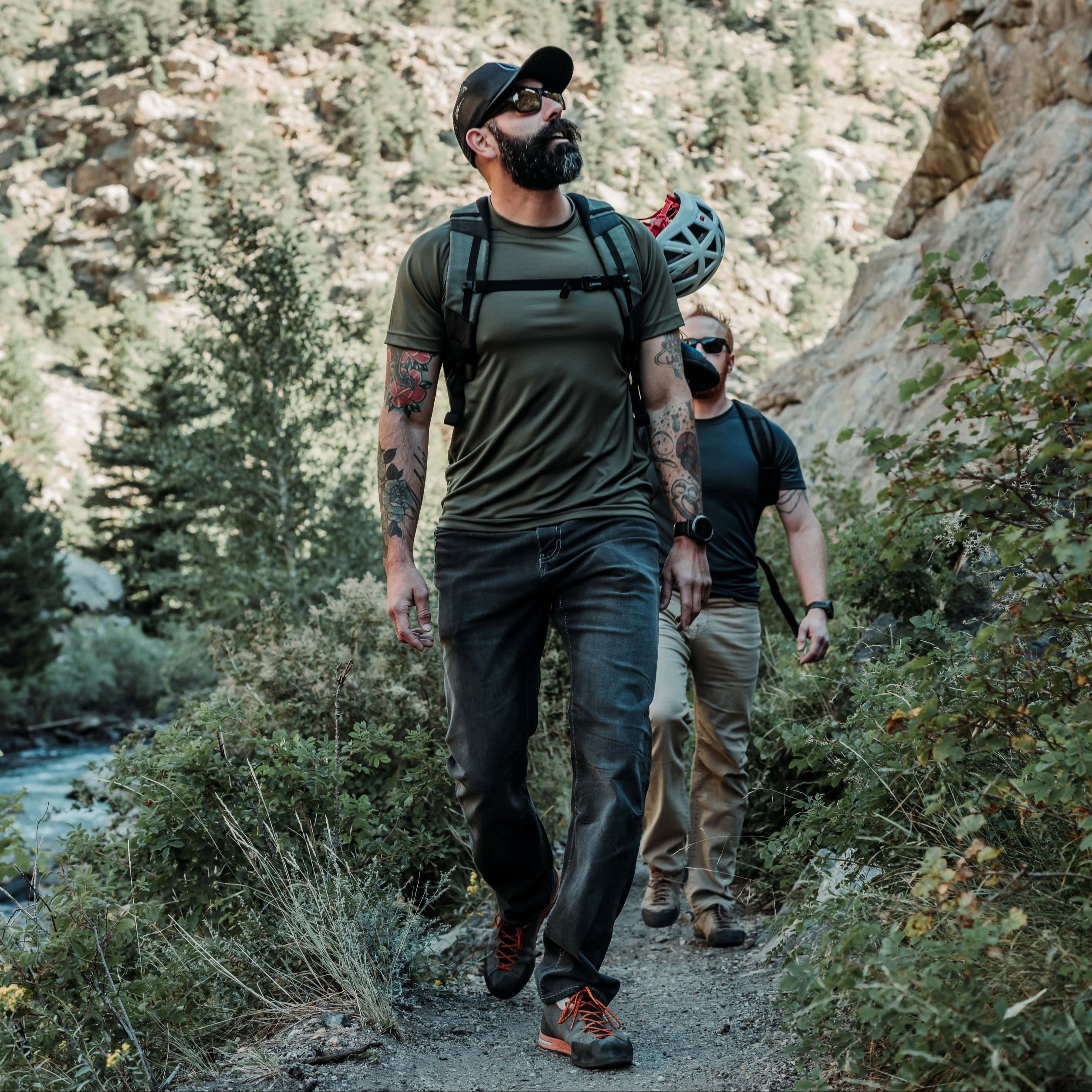 Photograph of two men hiking a trail near a river with rocky cliffs and lush green vegetation featuring a man with tattoos olive shirt and orange shoelaces
