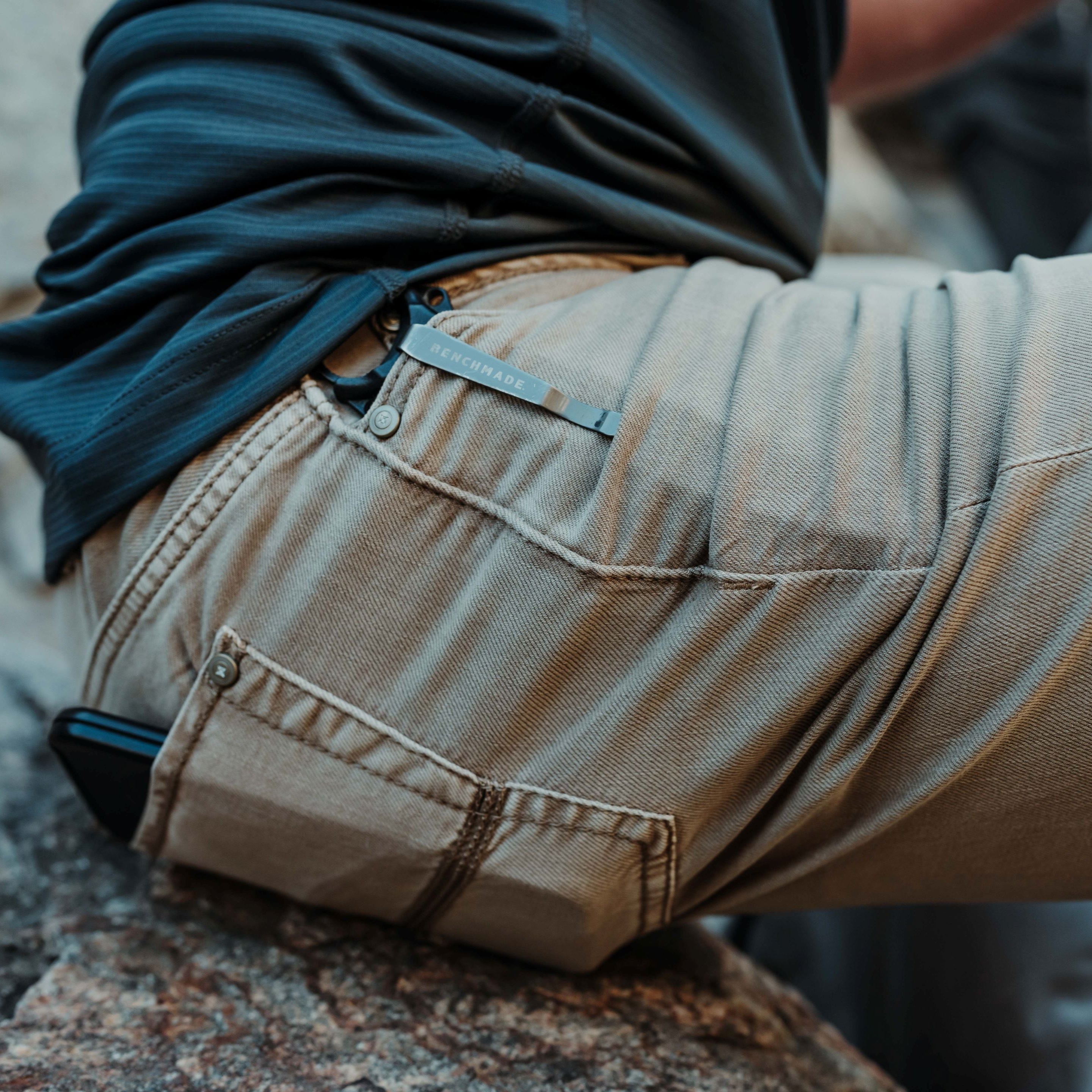 Photograph showing a person wearing khaki pants with a Benchmade tool clipped to the back pocket and a phone in the front pocket sitting on a textured rock outdoors