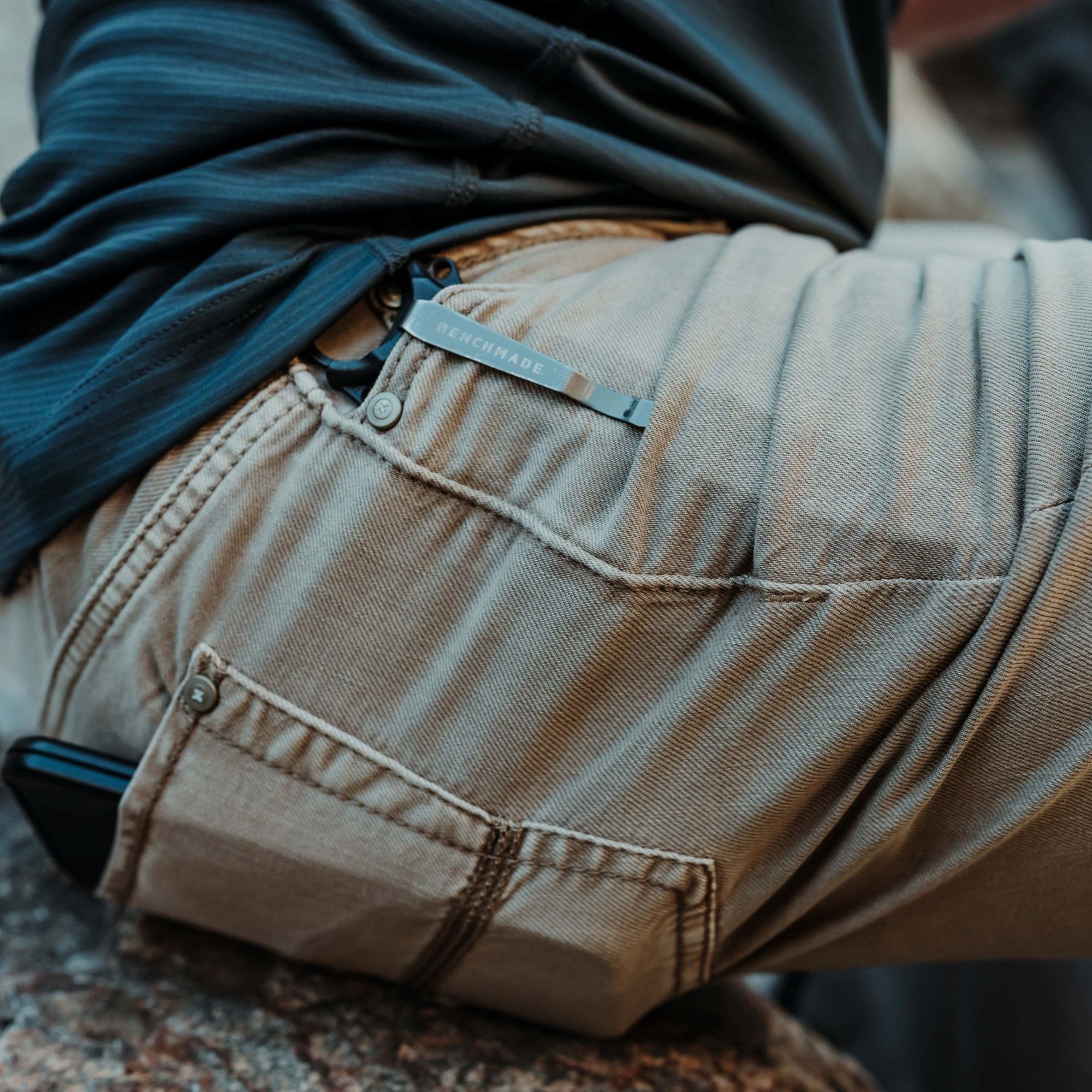 Photograph showing a person wearing khaki pants with a Benchmade tool clipped to the back pocket and a phone in the front pocket sitting on a textured rock outdoors