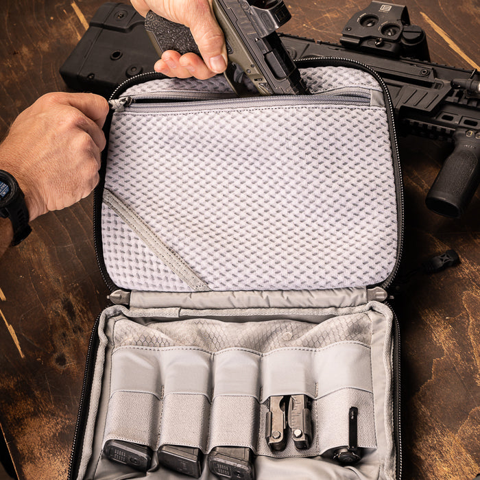 Photograph showing a person placing a handgun into a gray and black padded gun case containing magazines and a multi-tool on a wooden surface