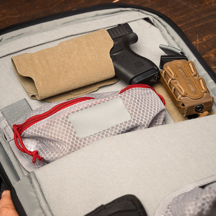 Photograph showing a Glock handgun in a tan holster inside a grey backpack along with a tan magazine pouch and a small red and grey mesh pouch a grey label and red zipper are visible
