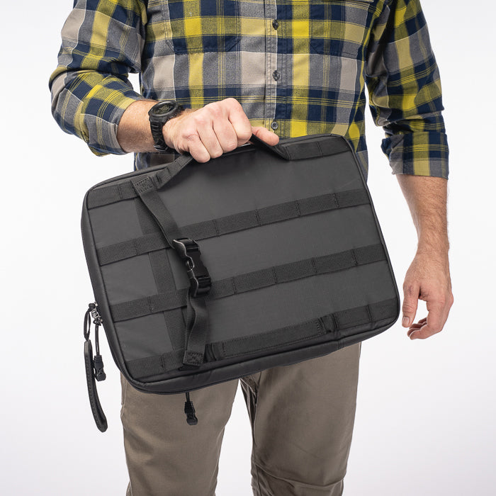 Photograph of a person holding a black laptop sleeve with adjustable straps and a handle featuring yellow and gray plaid shirt and a black watch