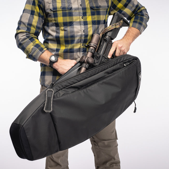 Product photography showing a person in a yellow and blue plaid shirt holding a black rifle case containing a rifle with scope and tactical accessories against a white background