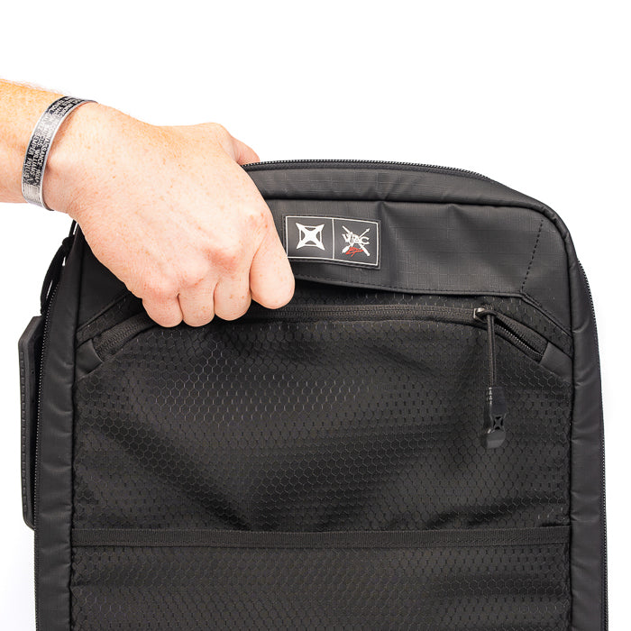Photograph of a black nylon bag with zippered compartments and a mesh pocket showing a hand opening the top featuring a logo and a branded patch with red accents