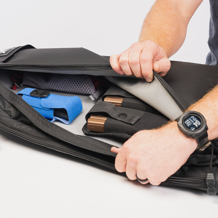 Product photography showing a black tactical range bag with gray interior containing two tan pistol magazines a blue accessory and other small items
