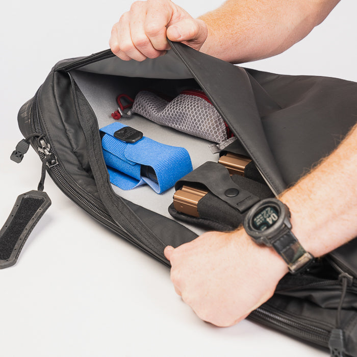 Product photography showing a black backpack's interior containing a blue pouch, tan magazine holders, and a gray mesh pocket with red accents
