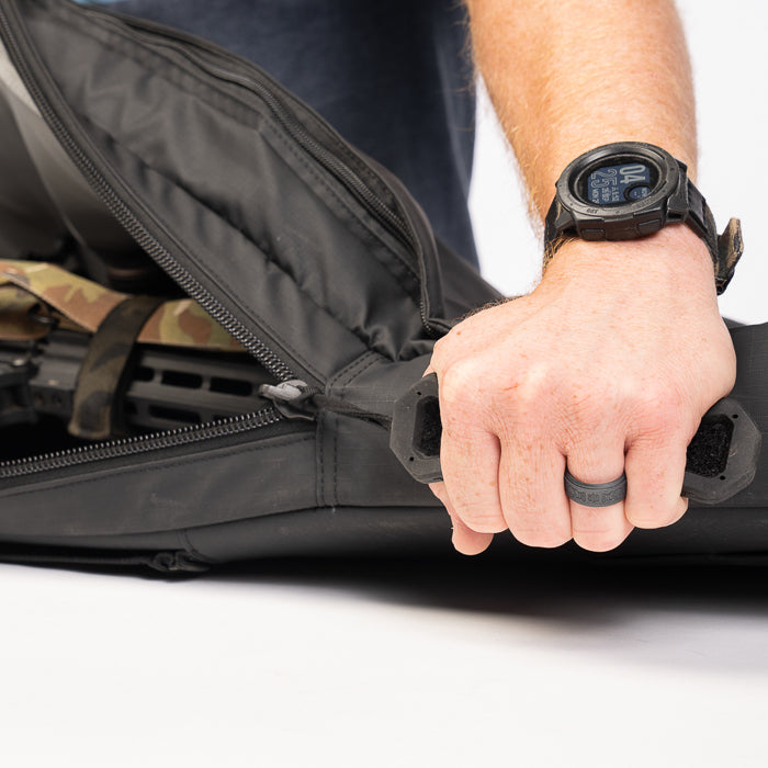 Product photography showing a person holding a black rifle case containing a partially visible firearm a camouflage-patterned accessory and a dark gray silicone ring on their finger with a black smartwatch visible on their wrist