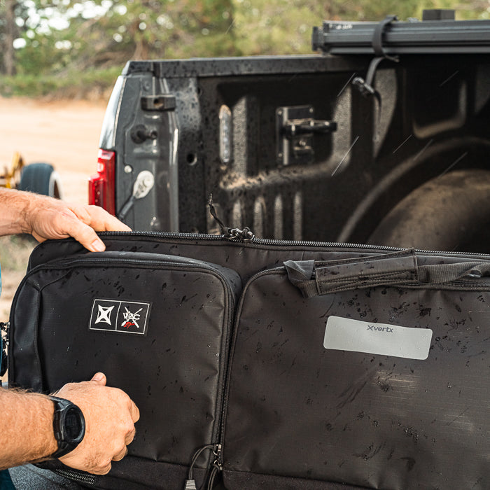 Photograph showing a person holding a black XVertx gun case with a silver logo patch near a gray pickup truck outdoors with rain drops on the truck bed