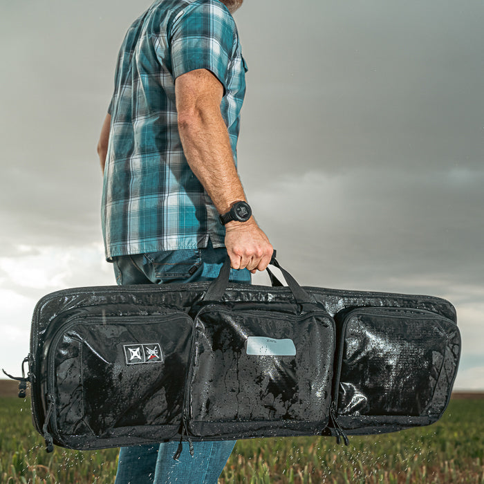 A photograph showing a man carrying a black, water-resistant rifle case in a grassy field under a stormy sky featuring a blue plaid shirt, a dark watch, and rain droplets on the case
