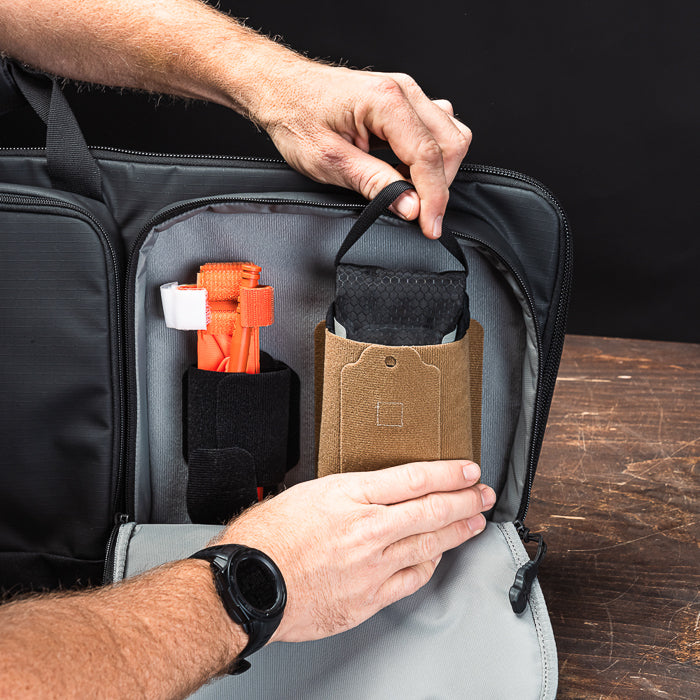 Photograph showing a person organizing the interior compartments of a black bag featuring an orange tool, a tan pouch with a small tag, and a black pouch with a textured surface