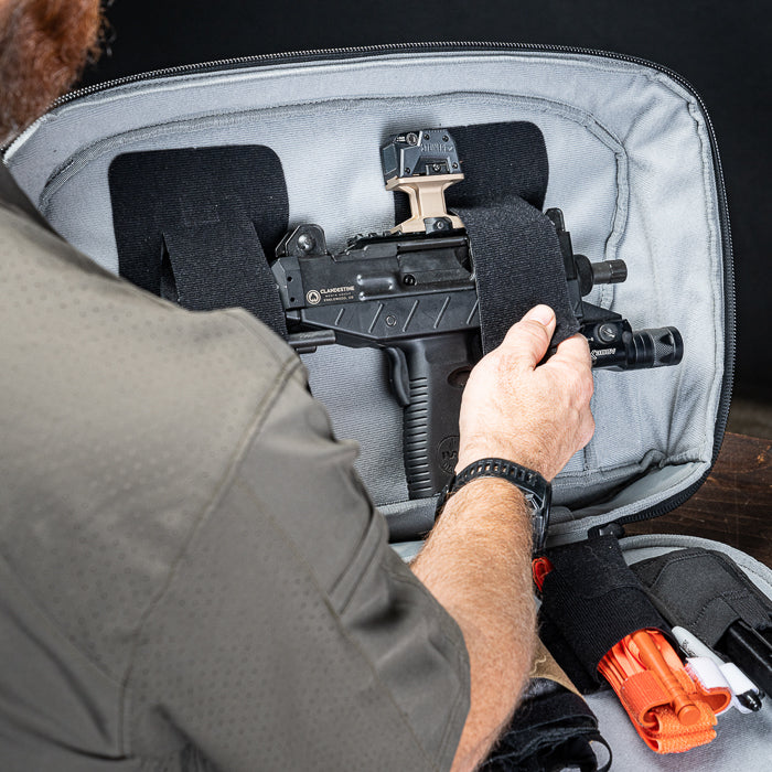 Product photography showing a person packing a clandestine firearms submachine gun with an attached tan optic and Surefire flashlight into a gray padded case also containing bright orange medical shears and white markers