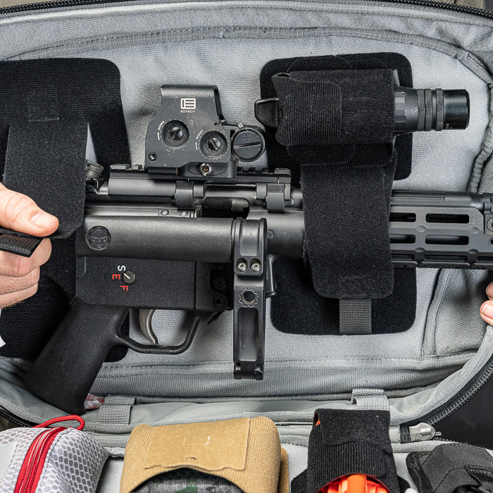 Photograph showing a black submachine gun with red markings and a holographic sight inside a grey backpack with black MOLLE straps and additional pouches