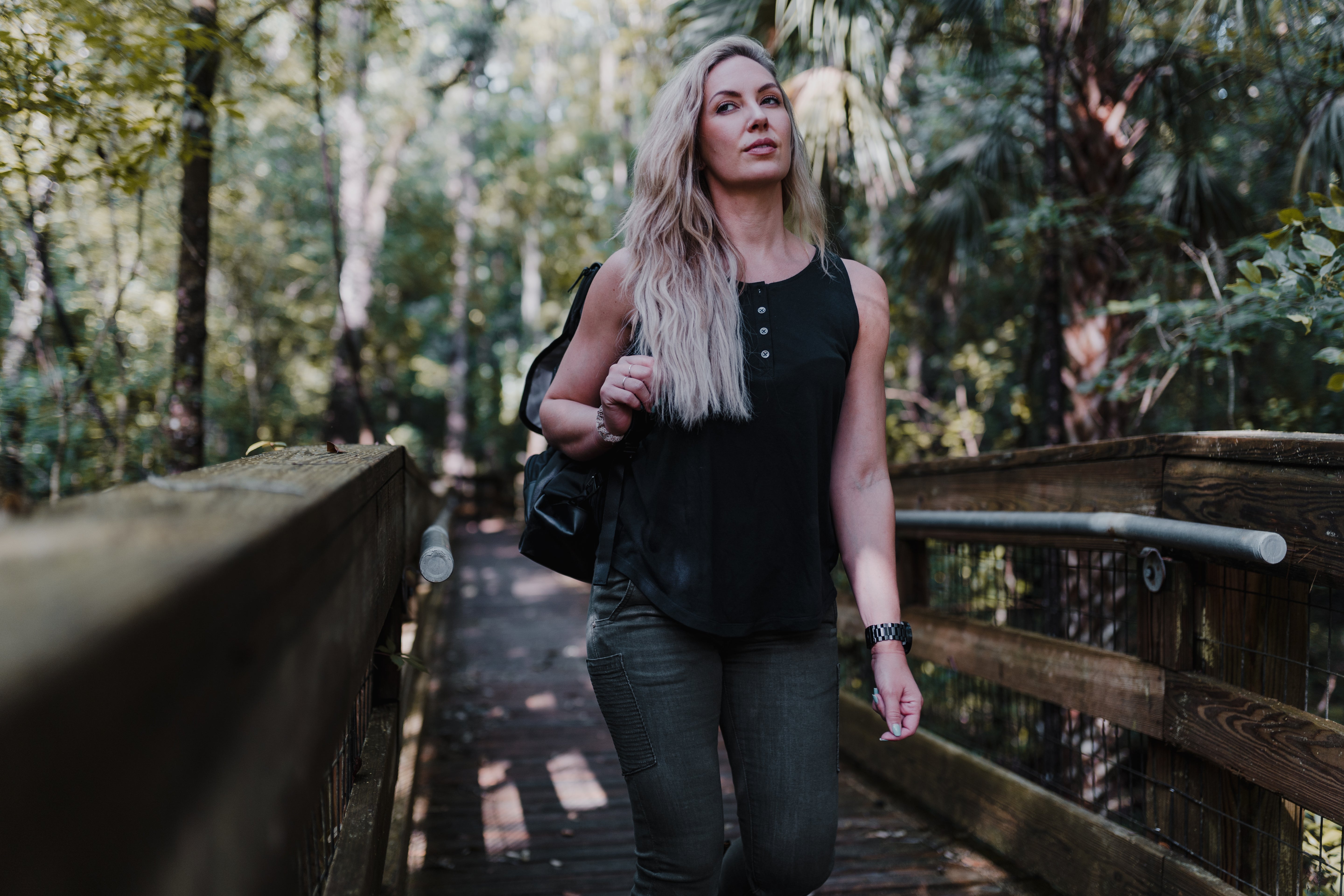 Photograph of a woman walking on a wooden boardwalk in a lush forest with palm trees and dappled sunlight filtering through the canopy, her black tank top, olive green cargo pants, and a black backpack creating a stylish yet practical look against the natural background
