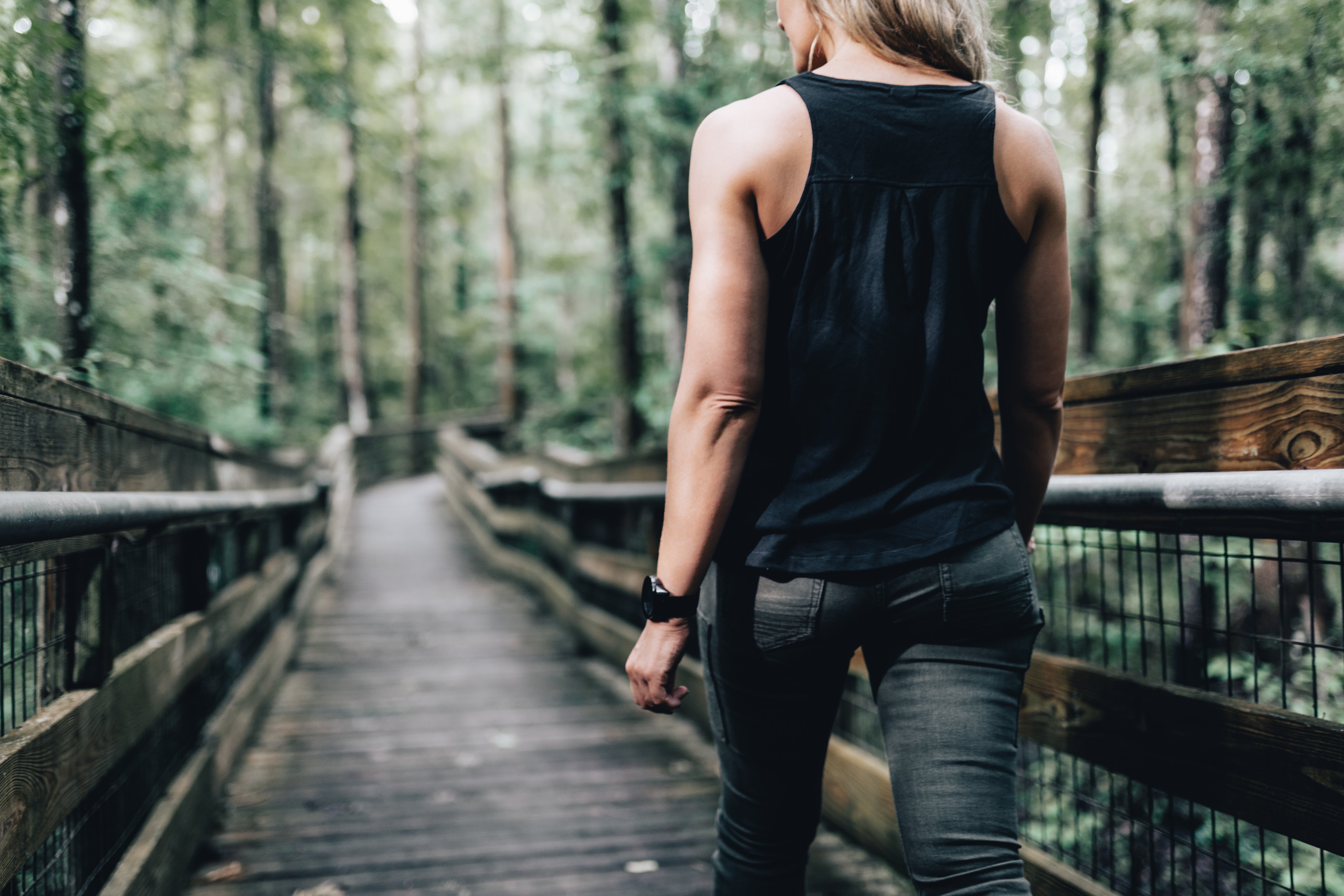 Photography of a woman in a black tank top and olive green jeans walking on a wooden boardwalk through a lush green forest with a dark smartwatch and visible wooden railings and metal mesh