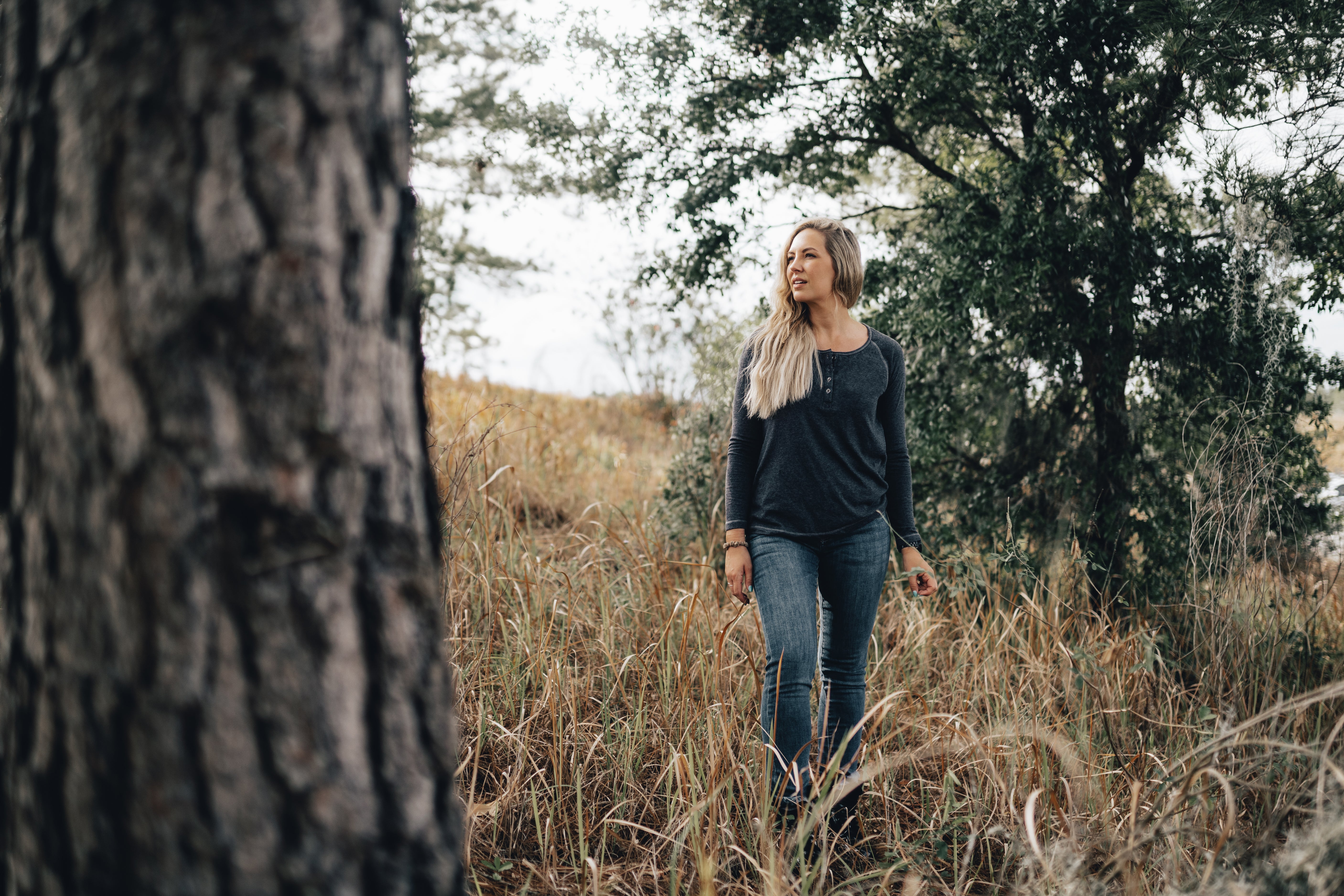 Photography A blonde woman walks through tall grass near a large tree and other greenery, showcasing her grey henley shirt, blue jeans, and the varied textures of nature's landscape
