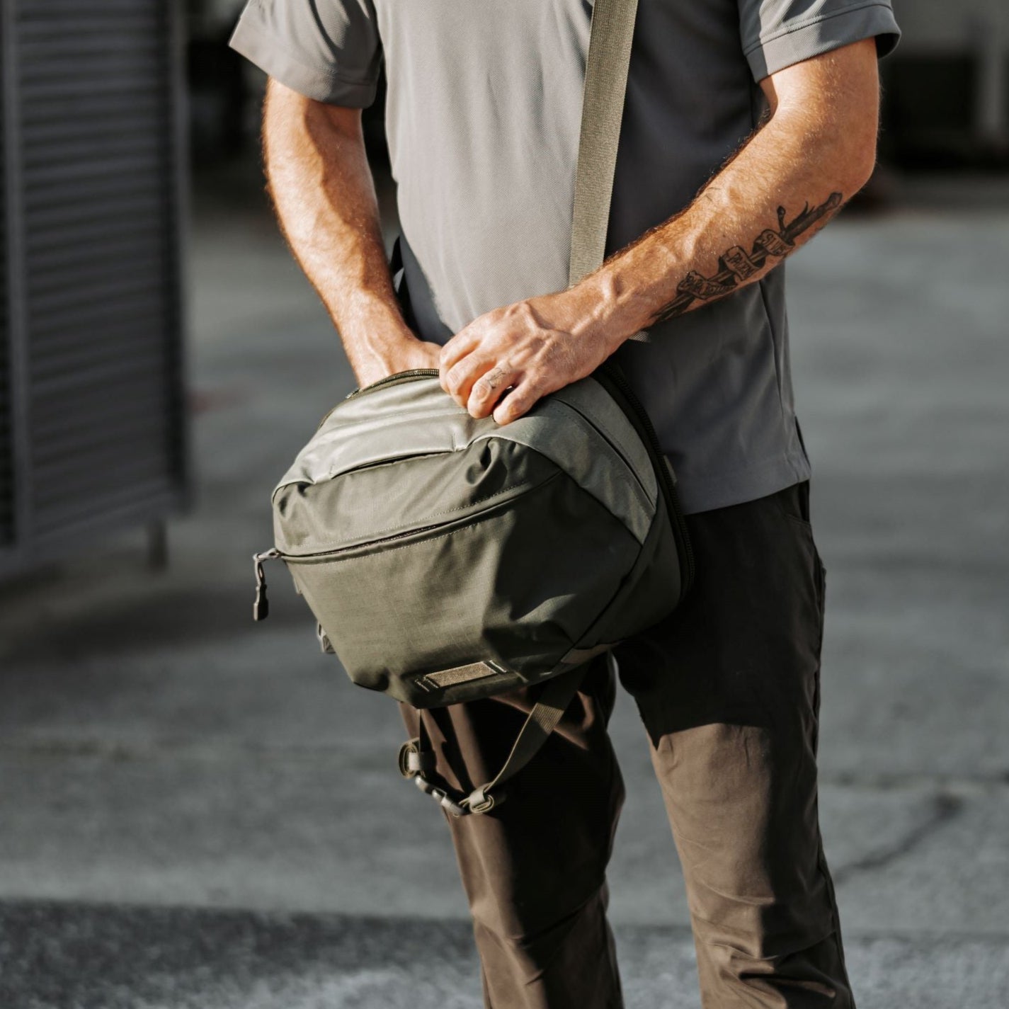 Photography unknown artist a man outdoors adjusting a muted olive-green crossbody bag with tan accents and a grey shoulder strap a grey shirt and brown pants visible and a detailed forearm tattoo
