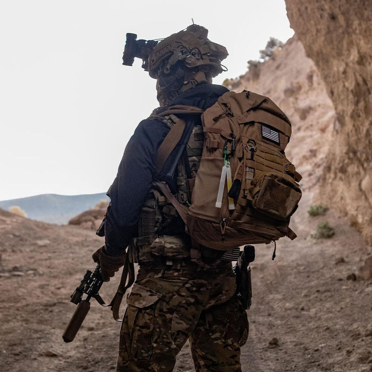 A person in full tactical military gear wearing a helmet with a night vision mount, a brown backpack featuring an American flag patch, and carrying a suppressed rifle stands in a rugged desert landscape near a rocky outcrop with glow sticks attached to their pack