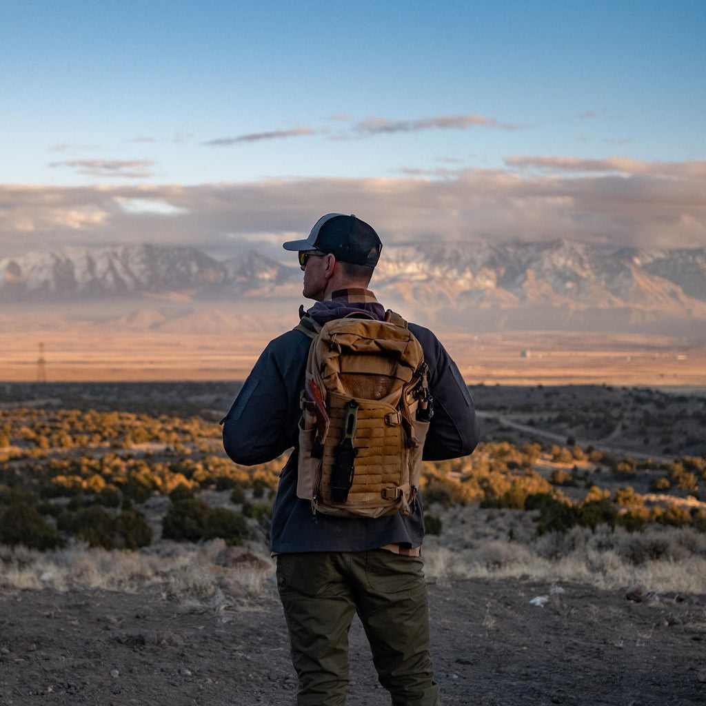 Photography of a man with a tan tactical backpack and utility knife, wearing a grey cap and sunglasses, observing a vast desert landscape with sparse golden scrub and distant snow-capped mountains under a vibrant blue and orange sky