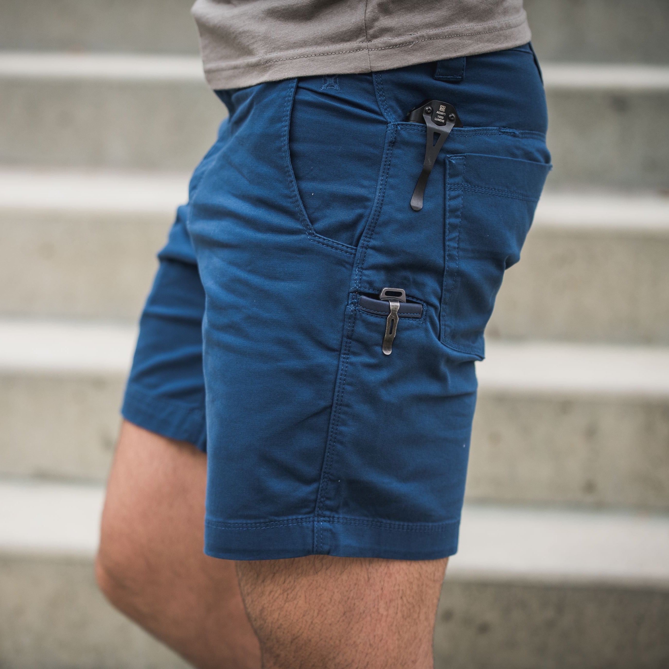 Photograph showing a man wearing dark blue shorts with utility pockets and a black multi-tool in a pocket against a blurry background of stone steps showcasing detailed stitching and dark metal hardware