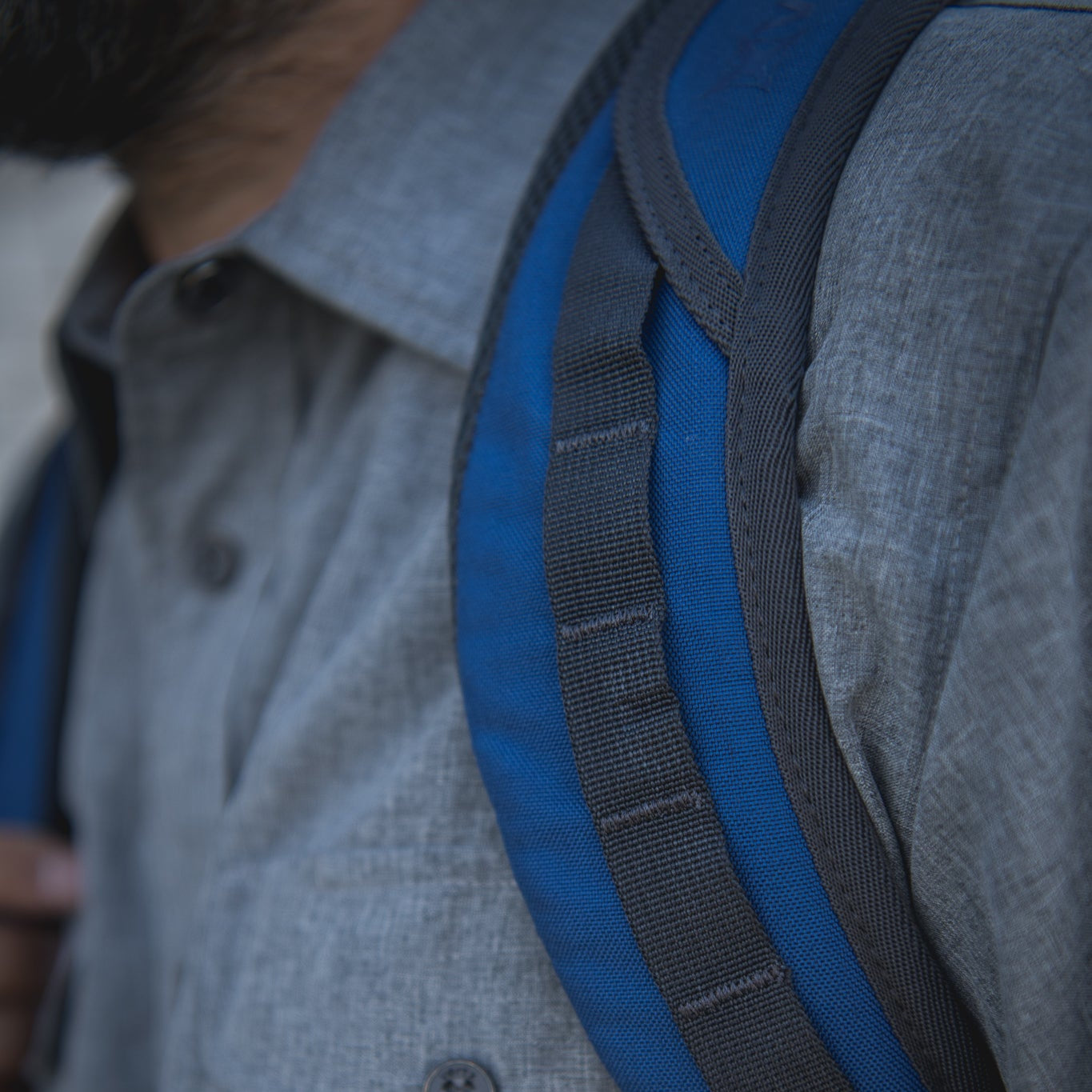 Photograph detail of a person wearing a gray shirt and a blue backpack with black straps and visible stitching