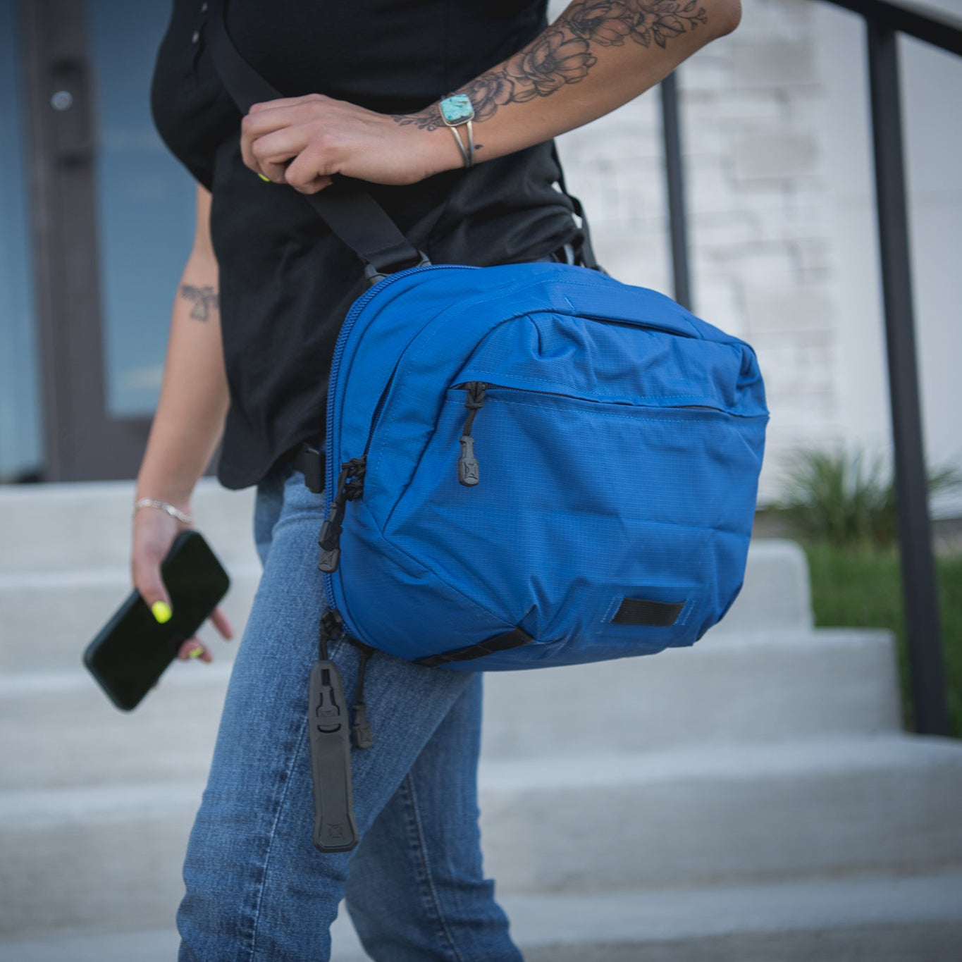 Photography model wearing a vibrant blue crossbody bag with black accents and zipper pulls near a modern building and steps featuring a turquoise bracelet and floral tattoos