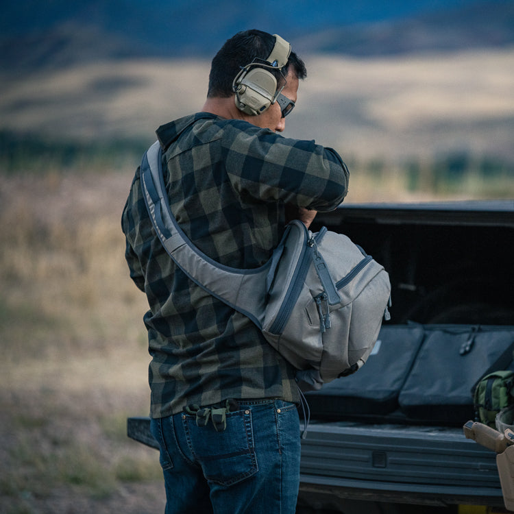 Photograph a man in a green and black plaid shirt packing a gray sling bag near a vehicle in a rural landscape featuring muted blue hills and tan grasses