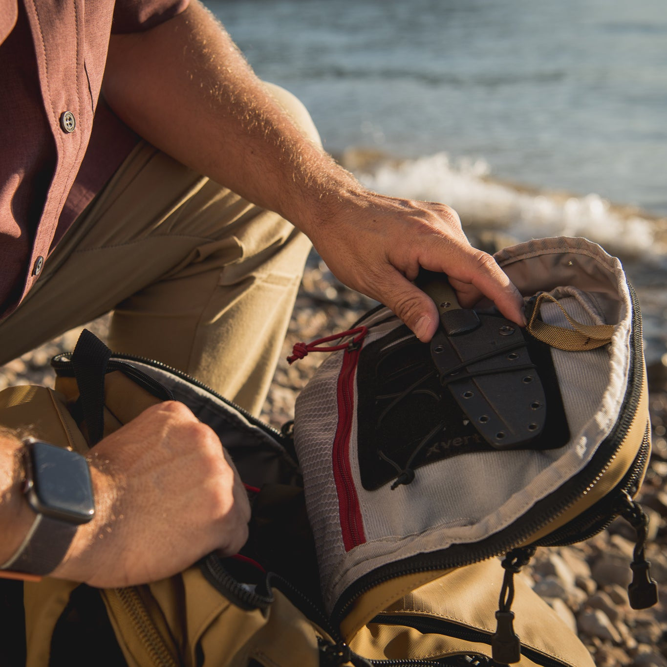 Photograph of a person placing a black knife with a tan handle into a tan backpack by a body of water with a sandy beach