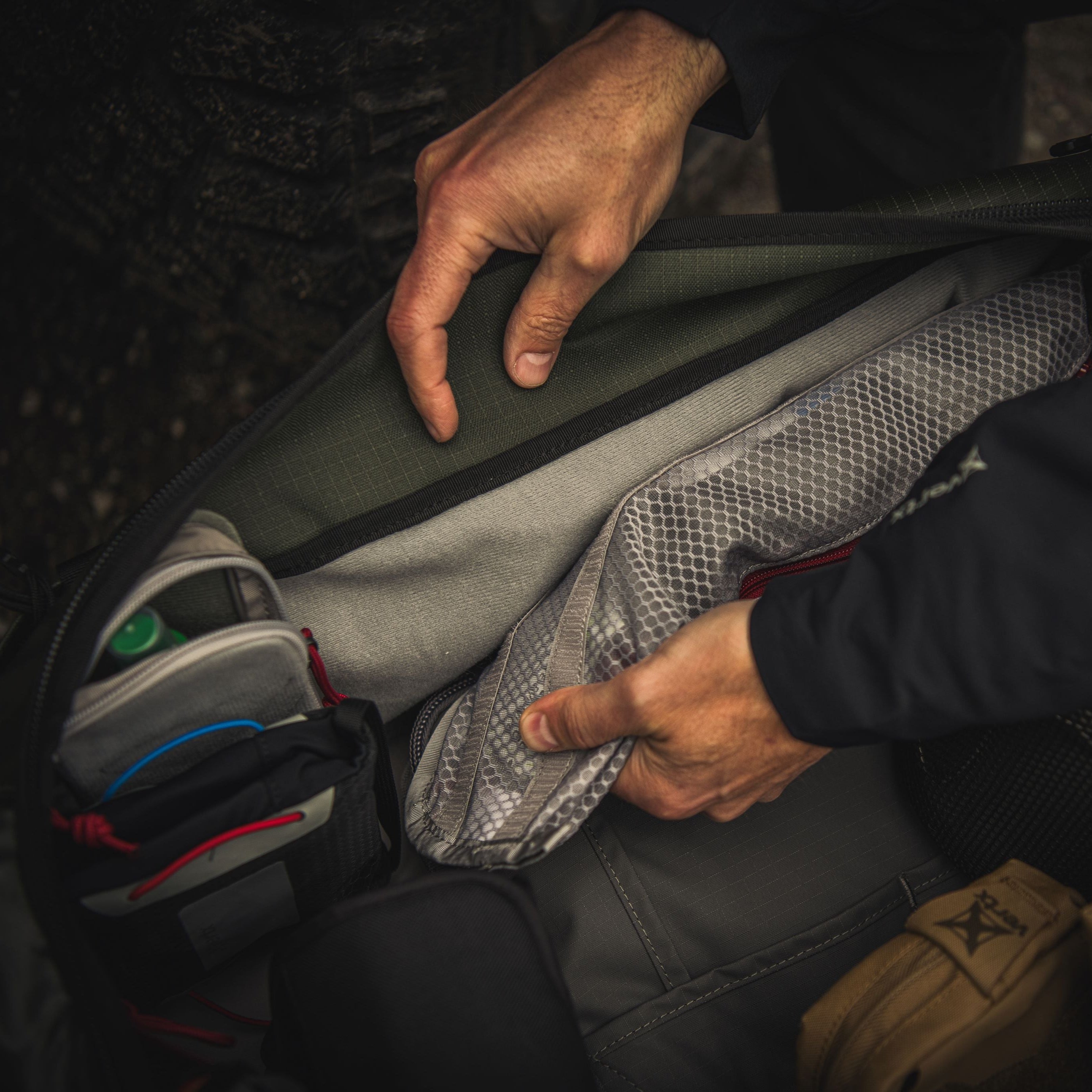 Photograph of a person unpacking a gray and olive green backpack featuring red accents a mesh pocket and a small zipped compartment