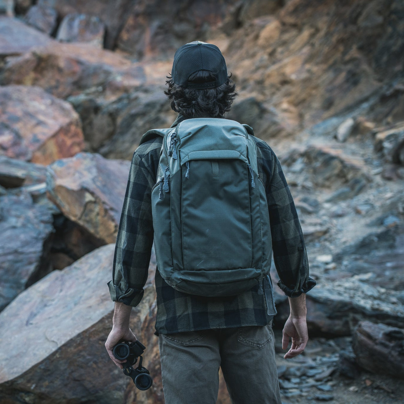 Photography a hiker with a green backpack and binoculars amidst a rocky landscape featuring large, rust-colored boulders and a textured, mountainous backdrop