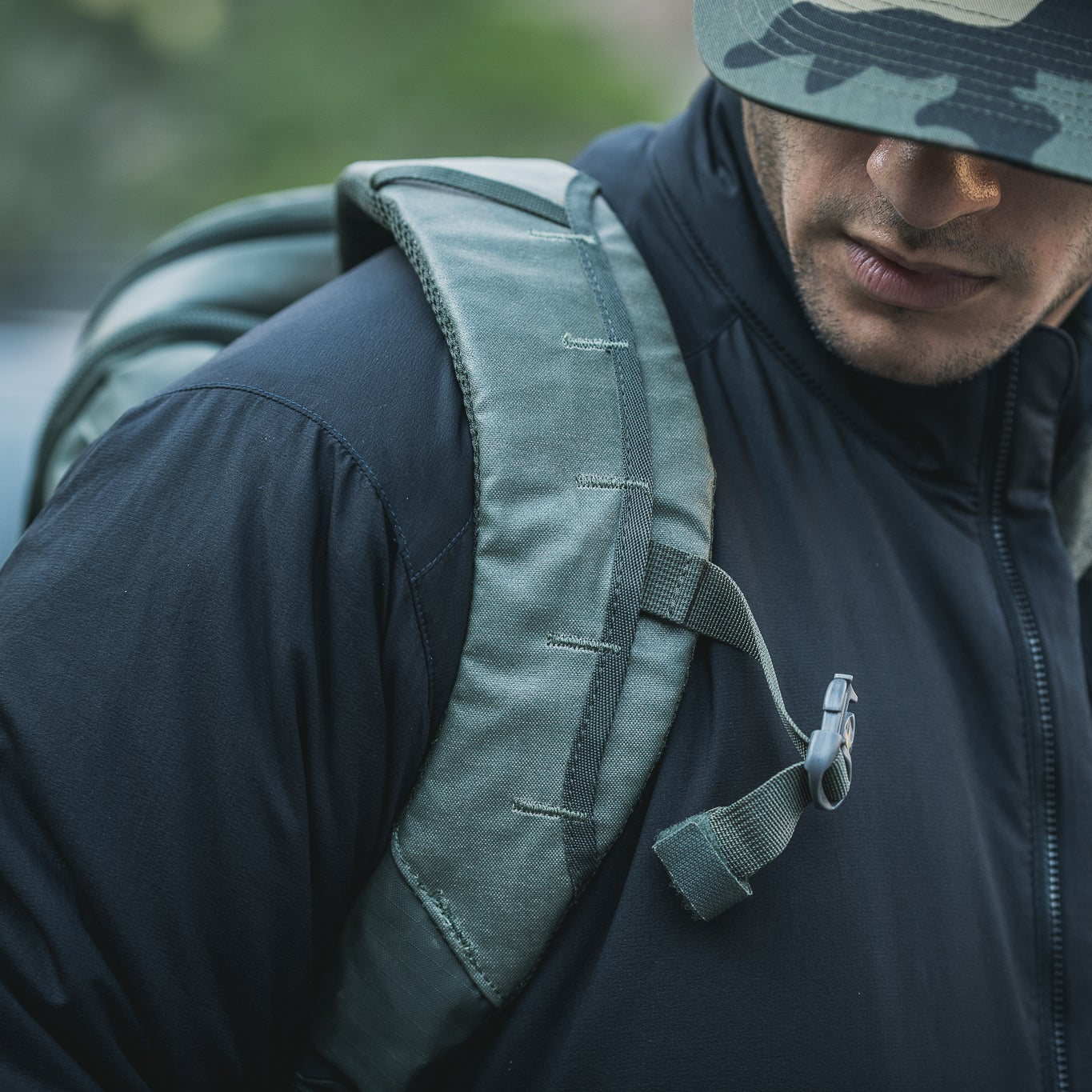 Photograph of a person wearing a dark jacket and camouflage hat carrying an olive-green backpack with visible straps and buckles outdoors