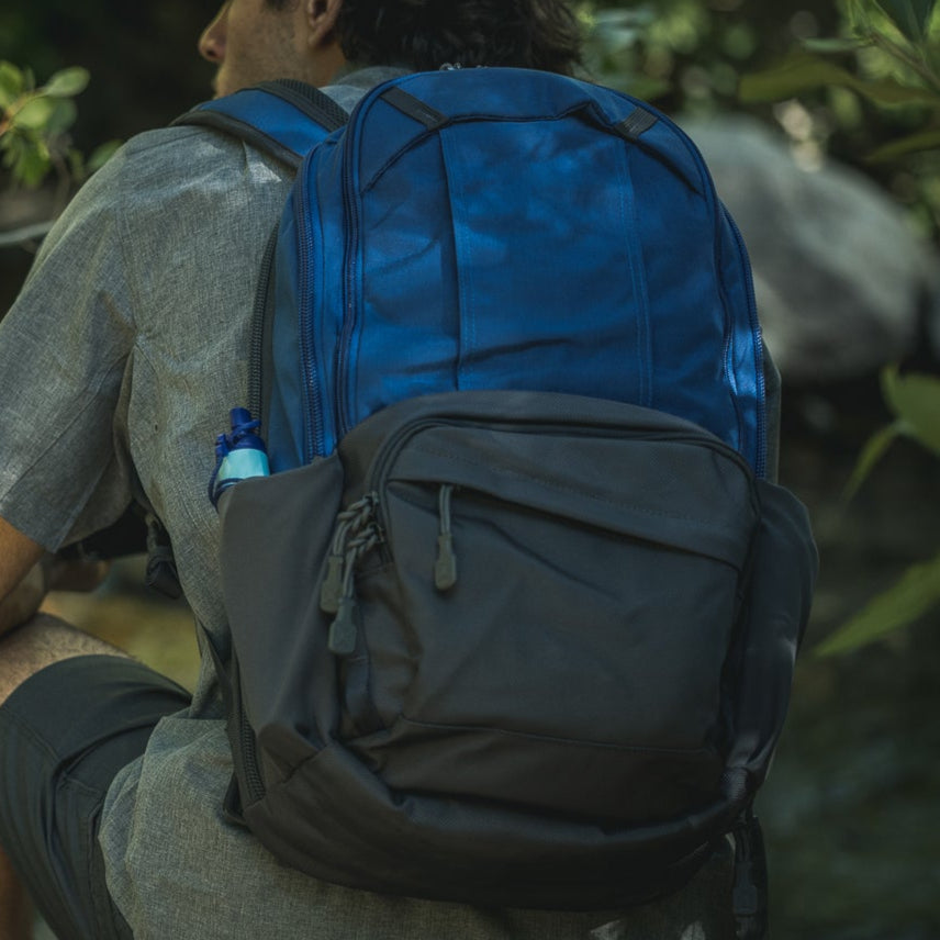 Photograph of a person wearing a blue and black backpack outdoors near lush green foliage and a rocky stream with visible adjustable straps and a small blue water bottle in a side pocket