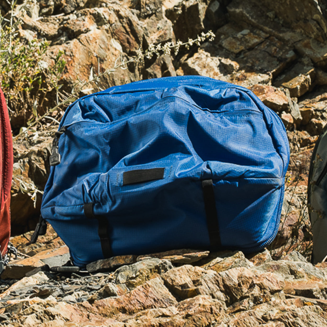 Photograph of a blue nylon backpack resting on rocks showing a black label, dark straps and a partially visible red backpack in the background
