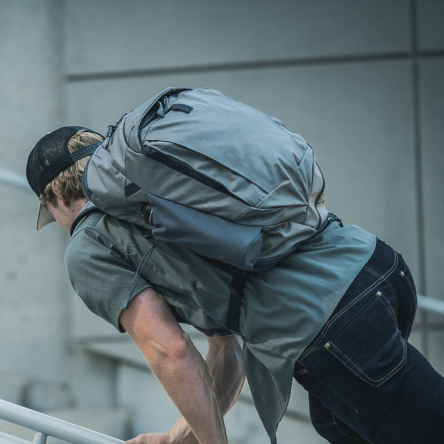 Photograph of a man descending a modern staircase wearing a gray backpack, olive shirt, and dark denim jeans, showcasing the bag's durable construction and sleek design against a muted urban backdrop