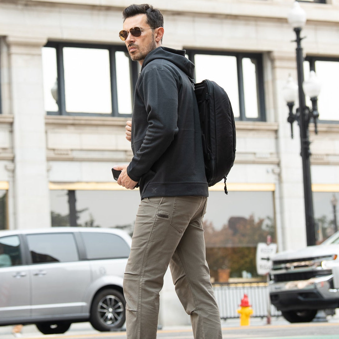 Photograph of a man in a black hoodie, olive green pants, and sunglasses walking on a city street with a black backpack, a bright yellow fire hydrant, and an ornate black street lamp in the background