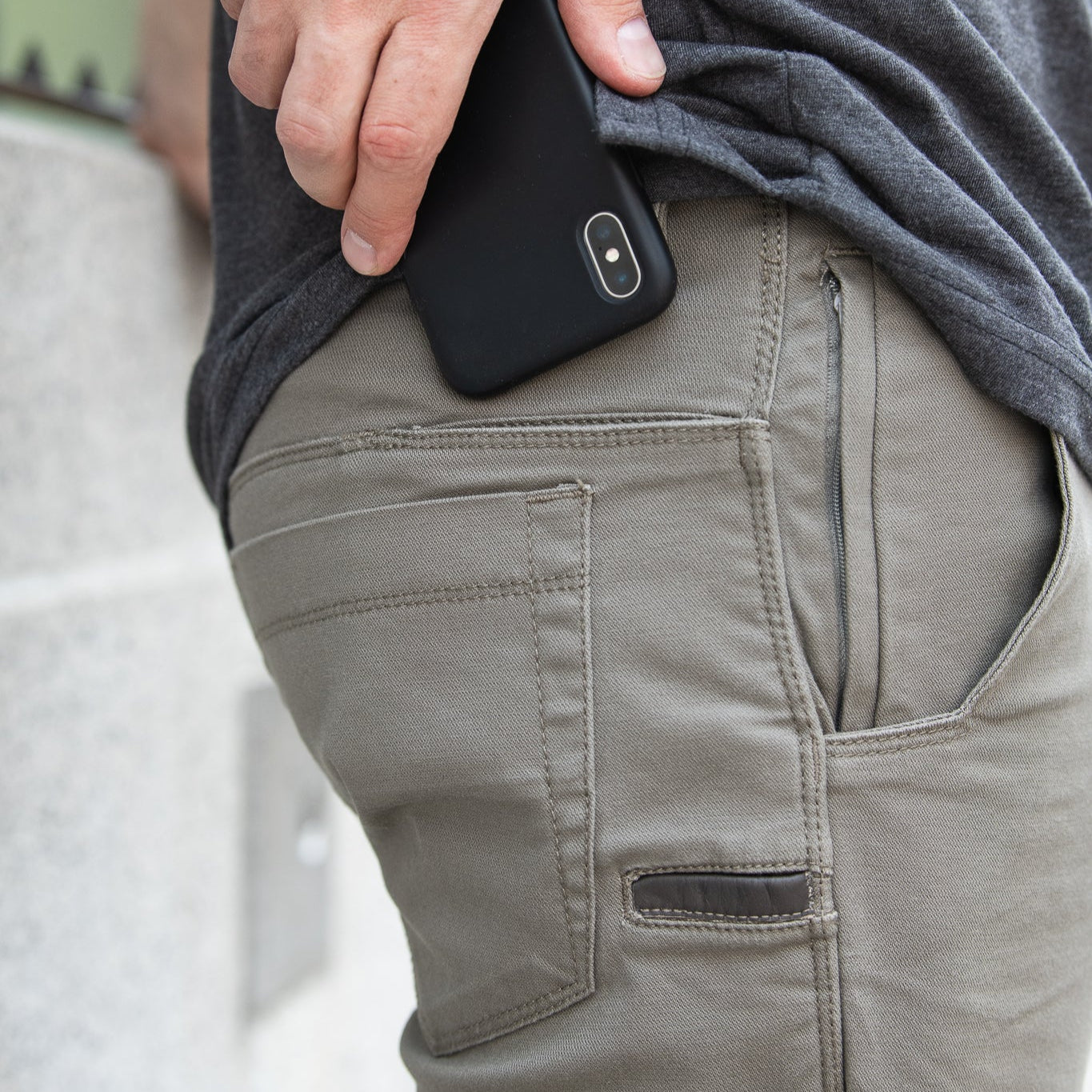 A person's hand placing a black smartphone into olive green tactical pants featuring a zippered side pocket and a dark leather accent on the front pocket