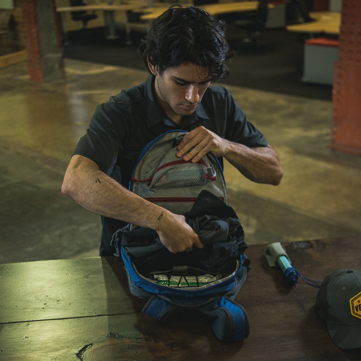 Photograph of a man packing a blue and gray backpack indoors featuring a water filter, energy bars, and a tattoo on his forearm