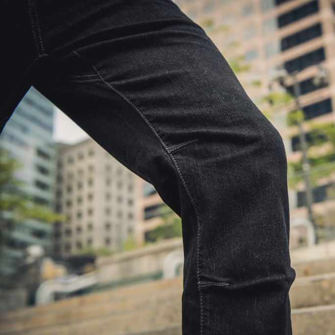 Photograph showing dark wash denim jeans with reinforced stitching and articulated knee detail against a blurry cityscape backdrop of modern high-rises and stone steps