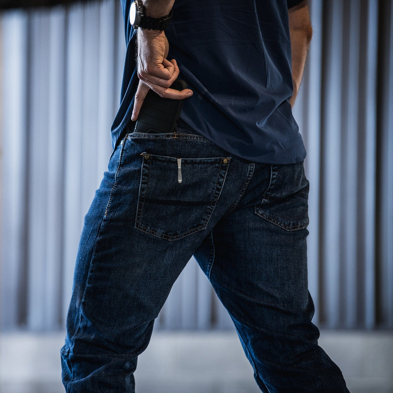 Photograph of a person in blue jeans and shirt placing a dark-colored object in their back pocket against a blurry light-grey background featuring subtle vertical lines and a hint of a white floor