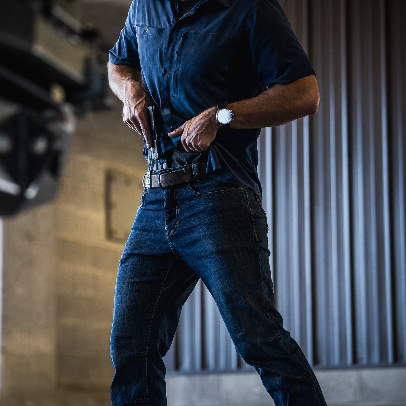 A photograph shows a man in blue shirt and jeans drawing a handgun from an IWB holster in an industrial setting with a gray corrugated metal wall and blurred background details