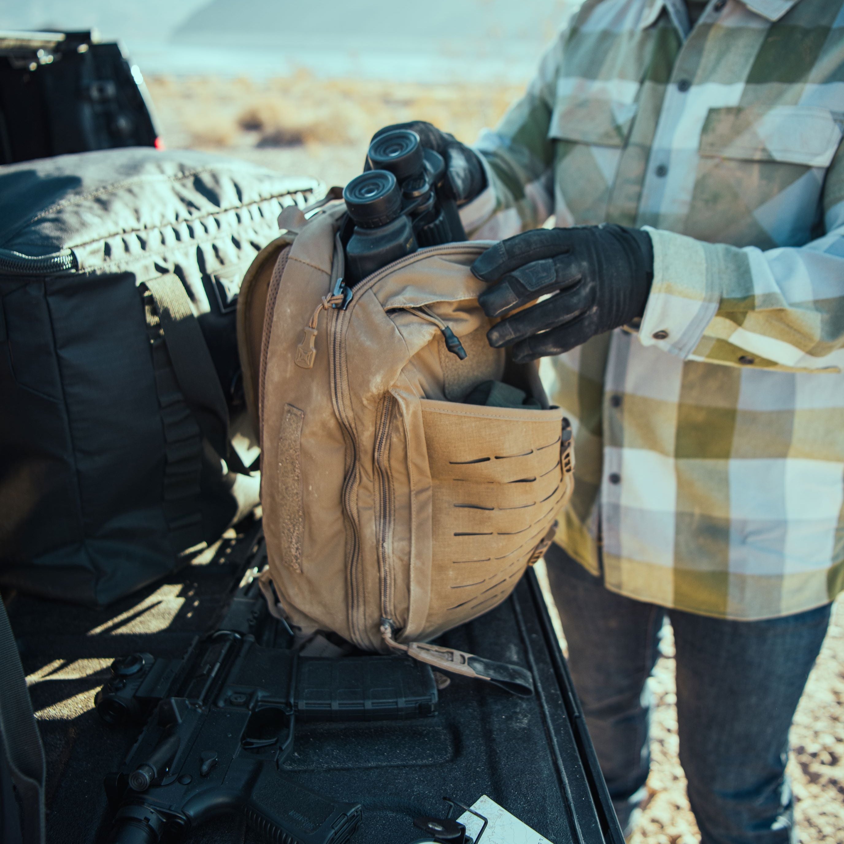 Photography of an individual in black gloves and a green checkered shirt organizing binoculars into a tan tactical backpack on a pickup truck tailgate with a rifle and map nearby in a vast desert landscape