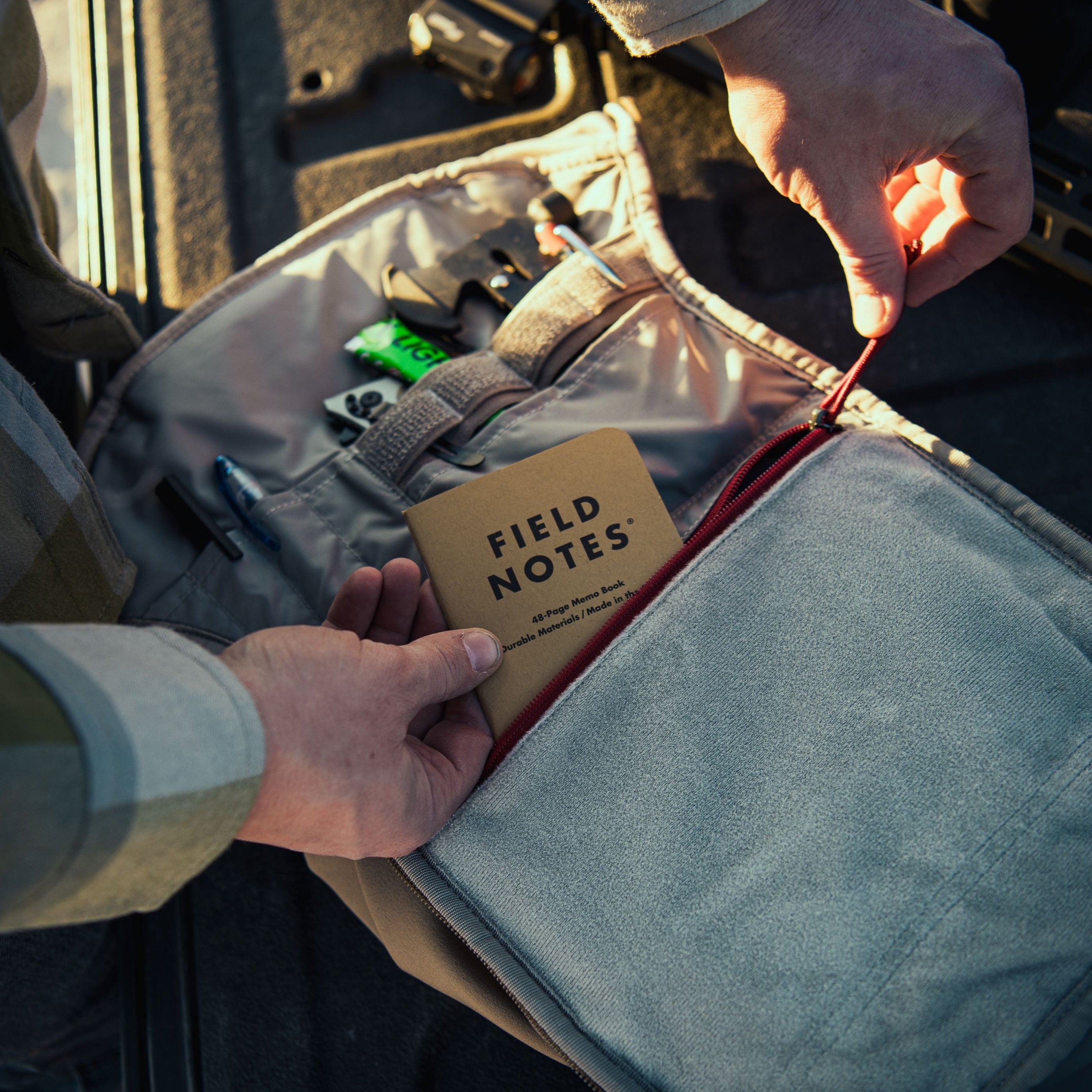Photograph of hands organizing a grey canvas field bag, placing a brown Field Notes memo book next to a vivid red zipper and a green chewing gum packet