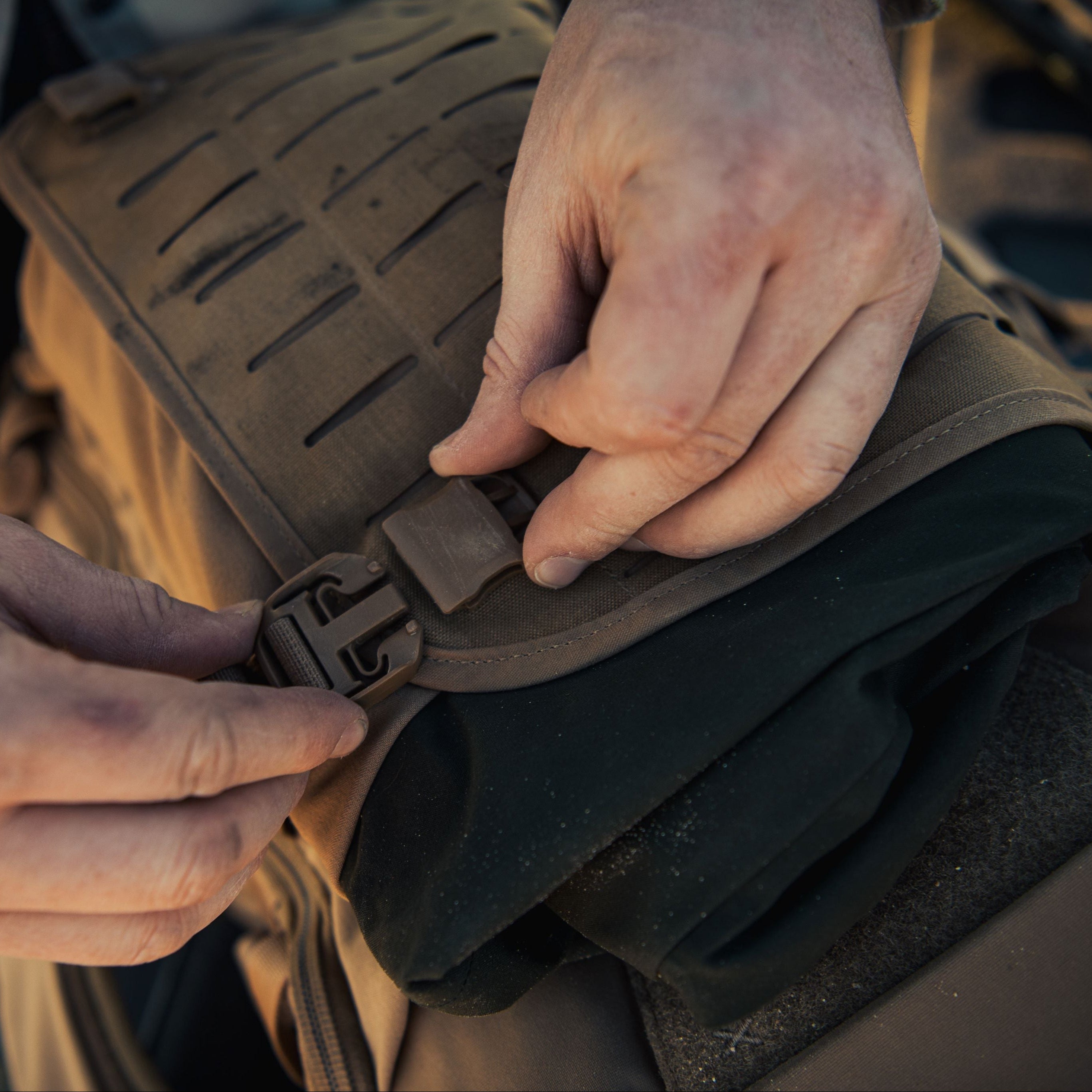 Close-up of dusty hands operating a tan plastic side-release buckle on rugged tan tactical gear featuring slotted webbing and a glimpse of dark green fabric